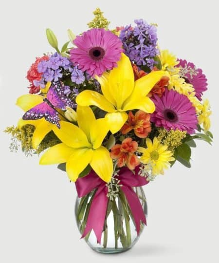 Brightly colored spring flowers, including lilies, gerbera daisies, and carnations, in a crystal clear glass vase. Tied with a coordinating bow and cute butterfly pick!
